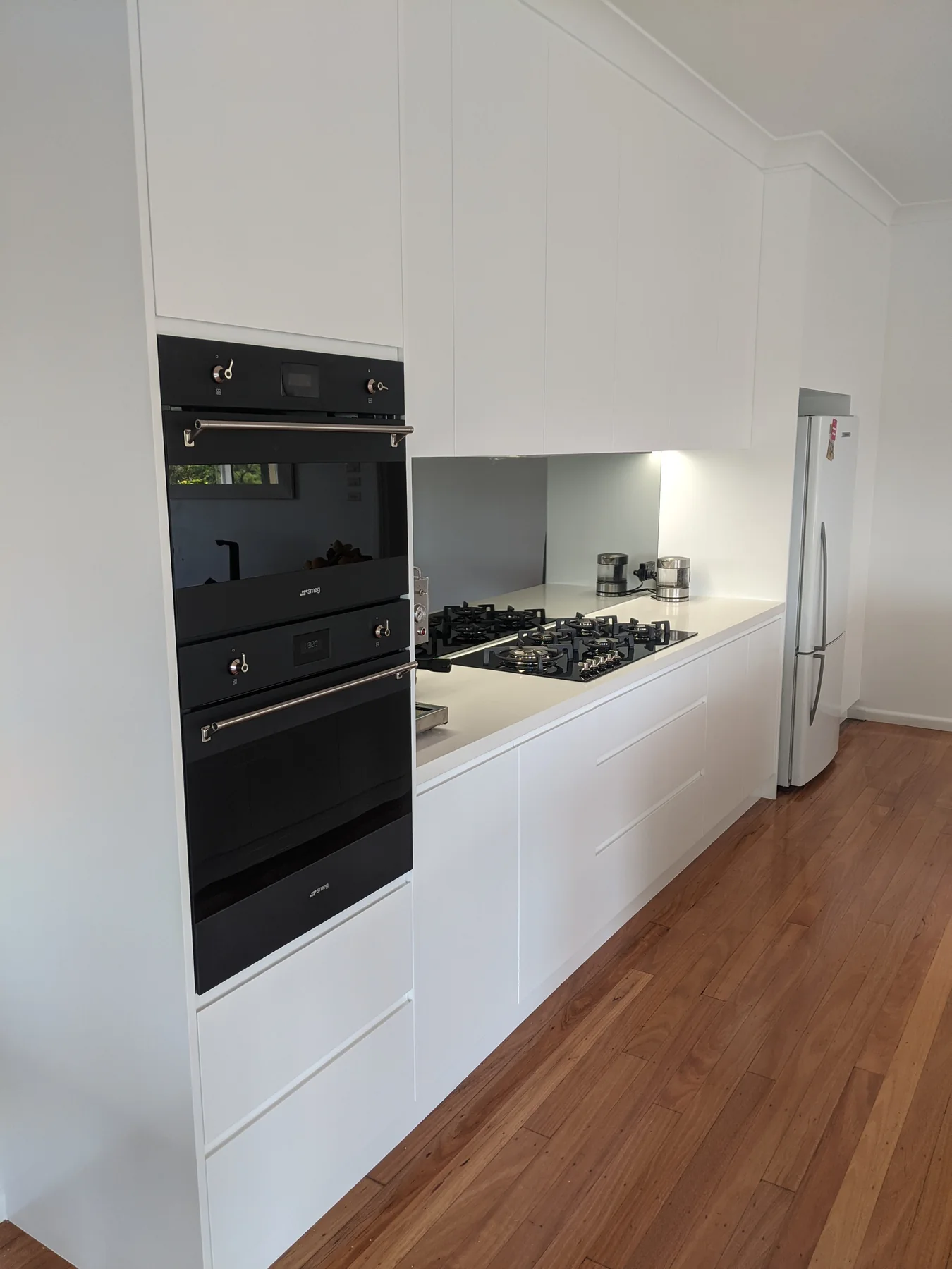 TCS kitchen with white cabinetry, stacked black ovens and timber flooring